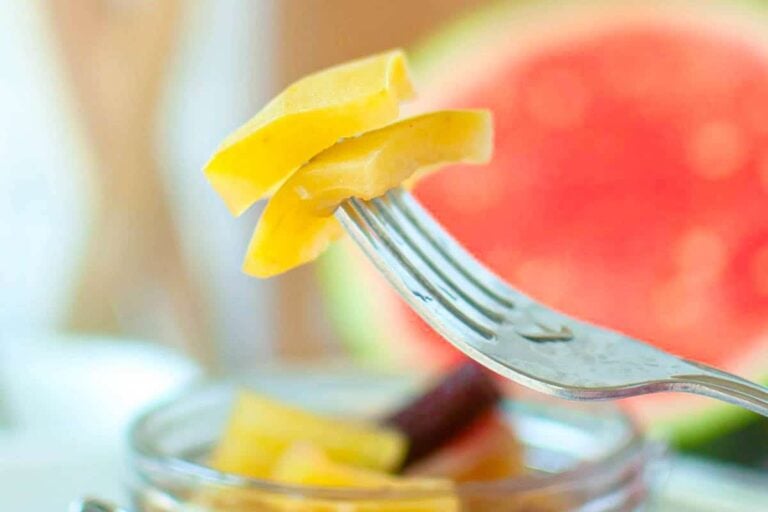 A close-up of a fork holding yellow pickled watermelon rind above a glass jar, with a blurred watermelon slice in the background.