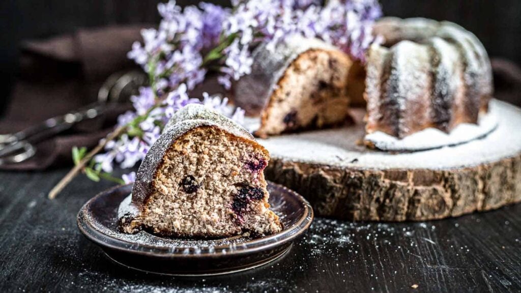 A slice of bundt cake with visible blueberries sits on a small plate, with the remaining cake and purple flowers in the background. The cake is dusted with powdered sugar.