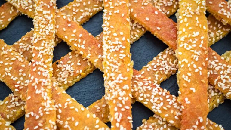 Close-up of several golden-brown sesame breadsticks arranged in a crisscross pattern on a dark surface.