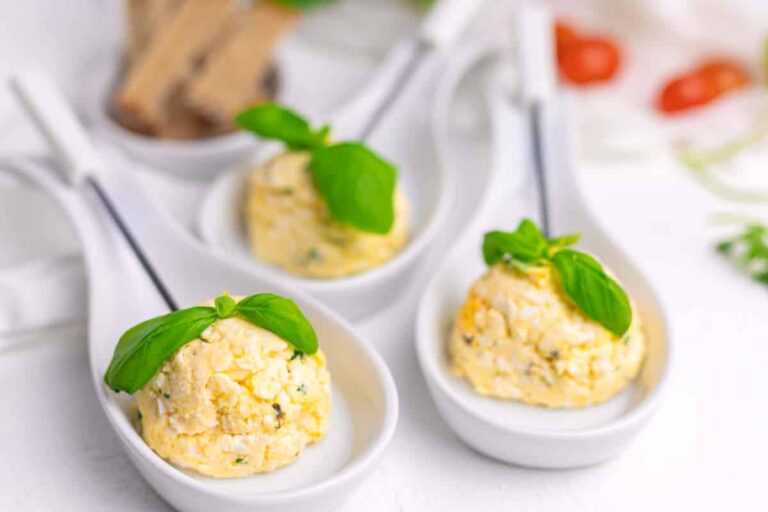 Three scoops of herbed cheese spread garnished with fresh basil leaves, each served on a white ceramic tasting spoon.