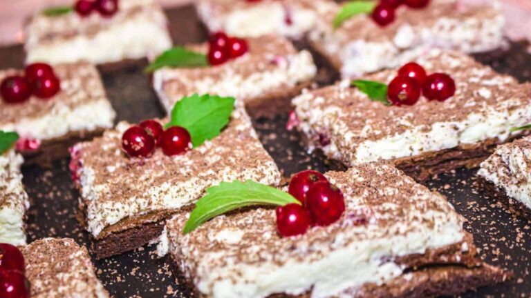 Rectangular dessert bars with layers of cream and chocolate, topped with red berries and green mint leaves, arranged on a dark serving tray.