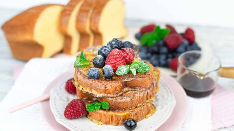 Three slices of French toast topped with fresh raspberries, blueberries, mint leaves, and powdered sugar on a plate, with sliced bread, berries, and syrup in the background.