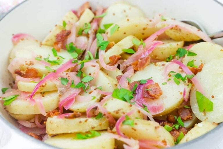 A bowl of sliced potatoes mixed with red onions, bacon pieces, and chopped parsley, served with a light dressing.