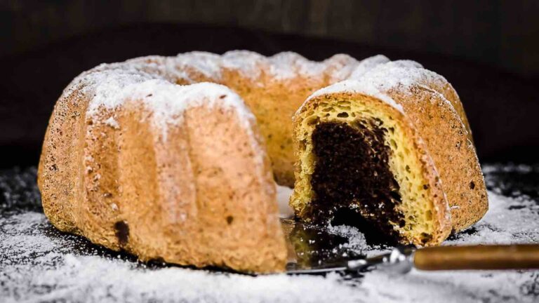 A marble bundt cake dusted with powdered sugar sits on a dark surface, with a single slice cut and resting on a cake server.