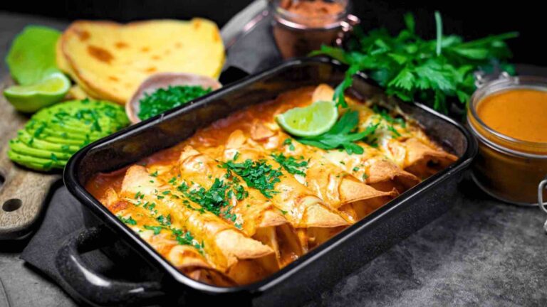 A baking dish of enchiladas topped with sauce and herbs, with sliced avocado, tortillas, lime wedges, and fresh greens on the side.