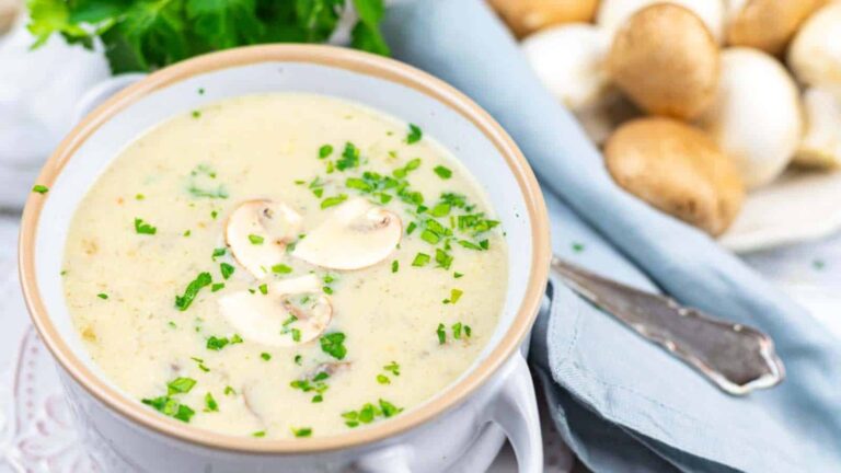 A bowl of creamy mushroom soup garnished with sliced mushrooms and chopped parsley, with whole mushrooms and a blue napkin in the background.