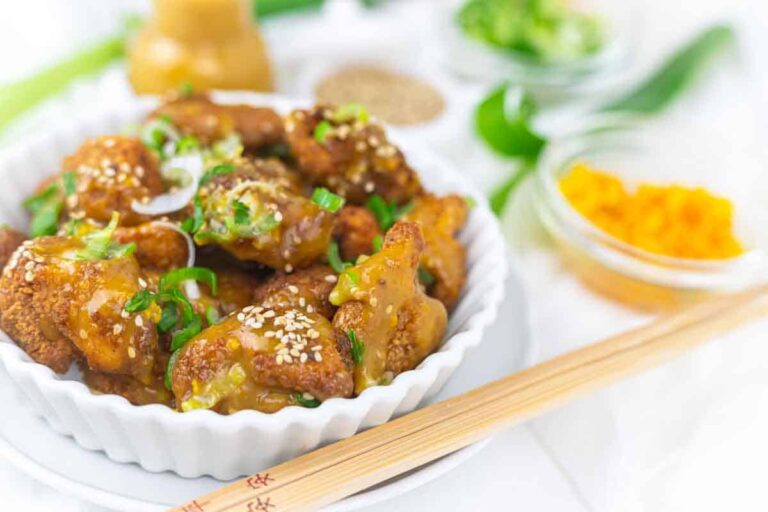 A white bowl of crispy glazed chicken garnished with sesame seeds and chopped green onions, with chopsticks and small bowls of various ingredients in the background.