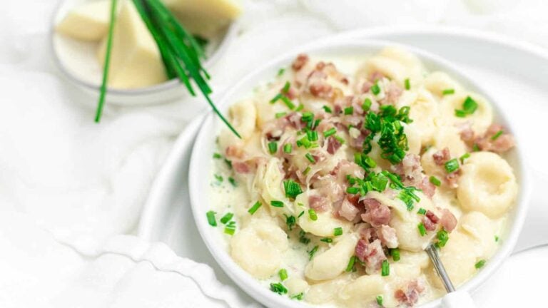 A bowl of creamy gnocchi topped with diced ham and chopped chives, with a fork; cheese wedges and whole chives are in the background.