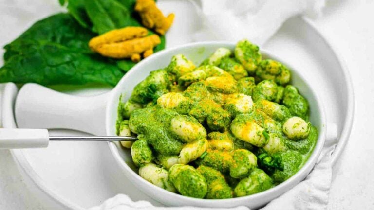 A white bowl of gnocchi covered in green pesto sauce, garnished with yellow powder, placed on a white plate with spinach leaves and yellow roots in the background.