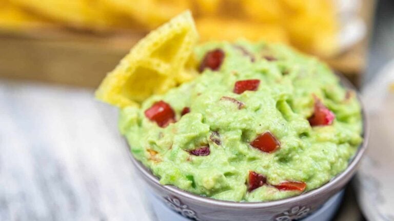 A bowl of guacamole with diced tomatoes and onion, garnished with two tortilla chips on the side.
