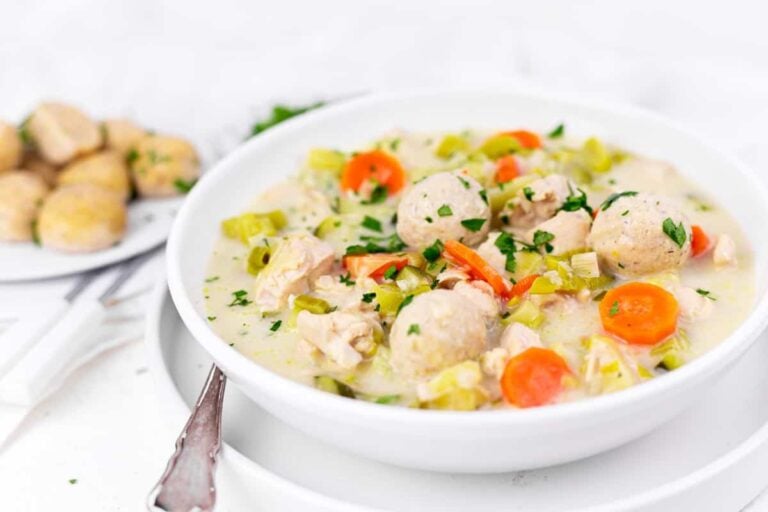 A bowl of chicken and vegetable soup with carrots, celery, and dumplings, garnished with fresh herbs. A plate of biscuits is in the background.