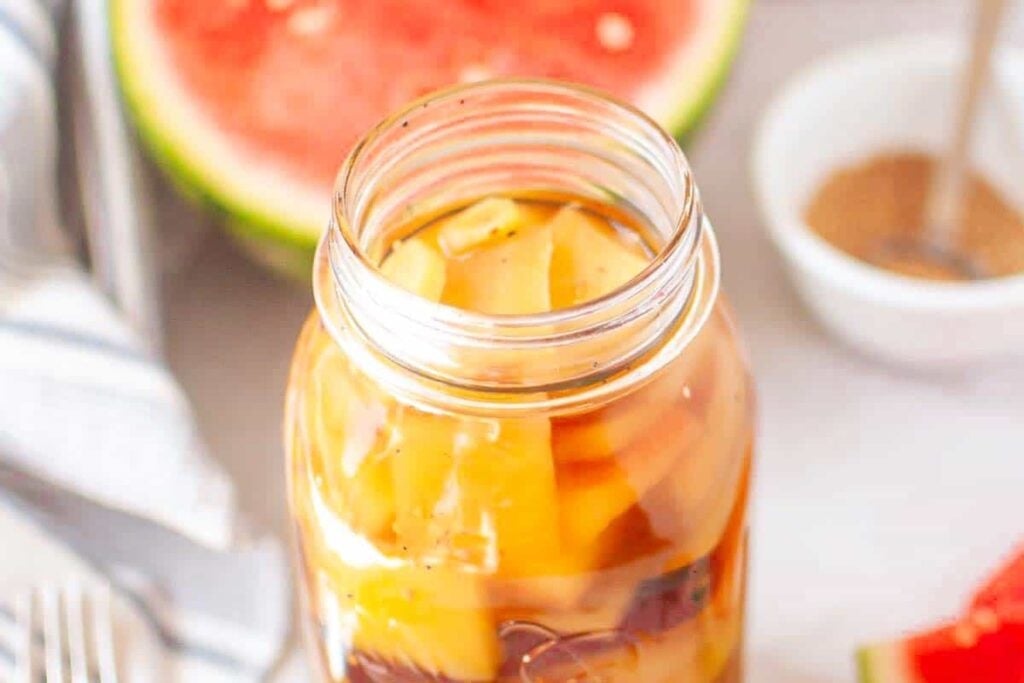 A glass jar filled with pickled watermelon rind, with a sliced watermelon and a small bowl of brown seasoning in the background.