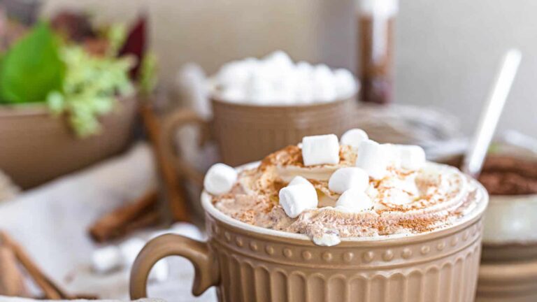 A close-up of a mug filled with hot chocolate, topped with whipped cream and mini marshmallows, with another mug and blurred background elements.
