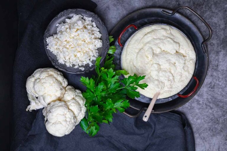 Overhead view of whole cauliflower, chopped cauliflower, fresh parsley, and a bowl of creamy cauliflower mash with a spoon on a dark surface.