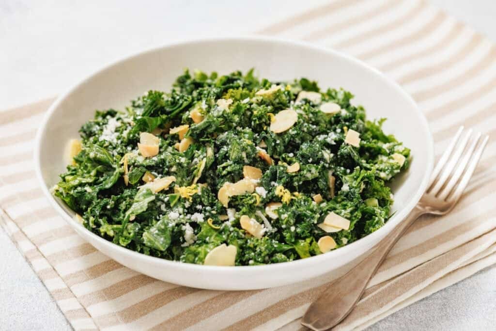 A white bowl of kale salad with lemon sits on a striped placemat beside a fork.