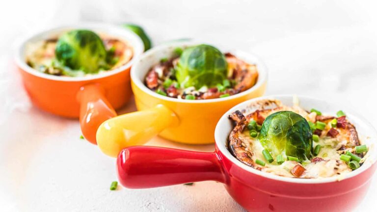 Three small casserole dishes with handles, each filled with a baked dish topped with a whole Brussels sprout and chopped green onions.