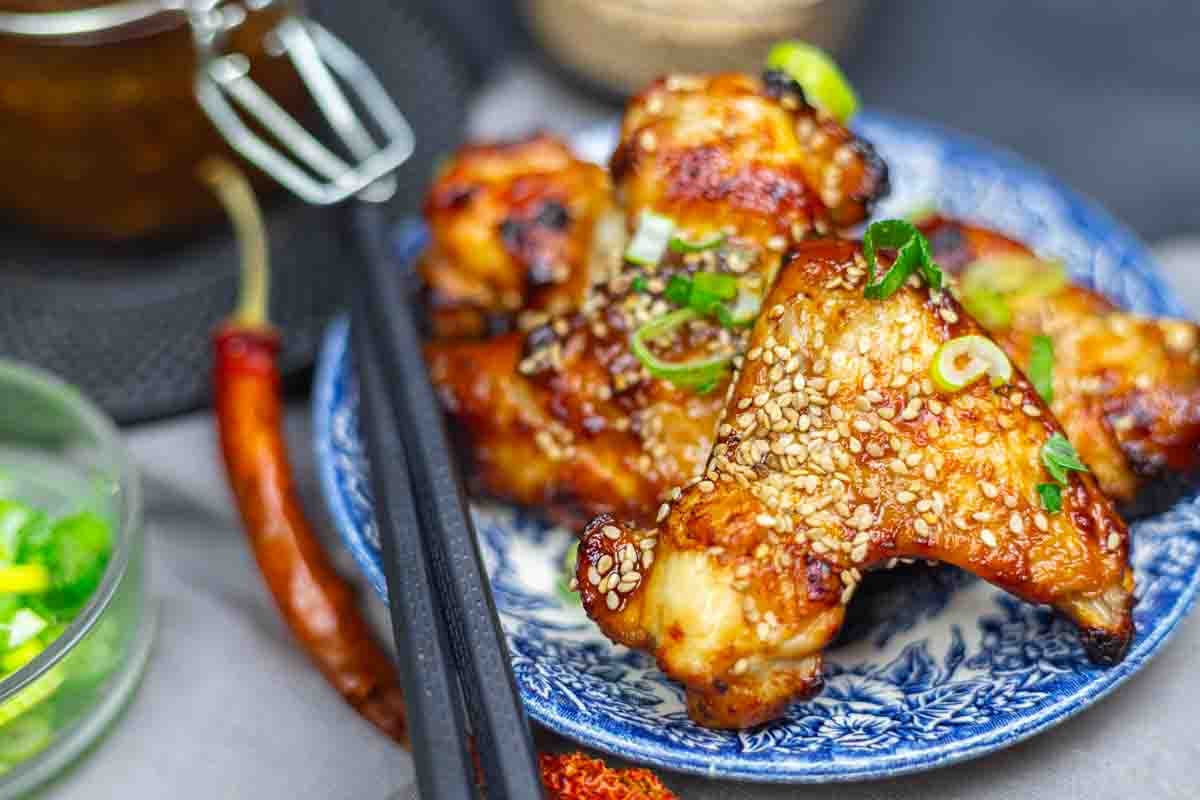 Grilled chicken wings glazed with sauce, topped with sesame seeds and green onions, served on a patterned blue plate with black chopsticks.