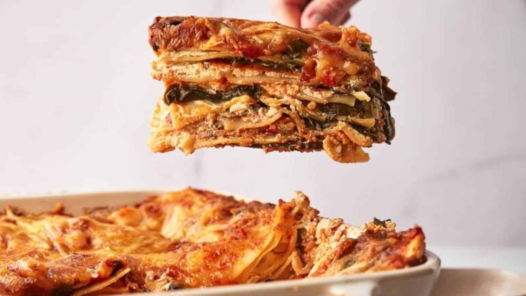 A hand lifts a square slice of vegetable lasagna from a baking dish filled with more lasagna.