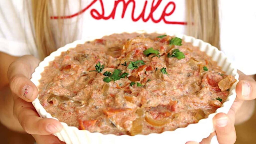 A person holds a white dish filled with a chunky, creamy tomato-based dip garnished with chopped herbs.