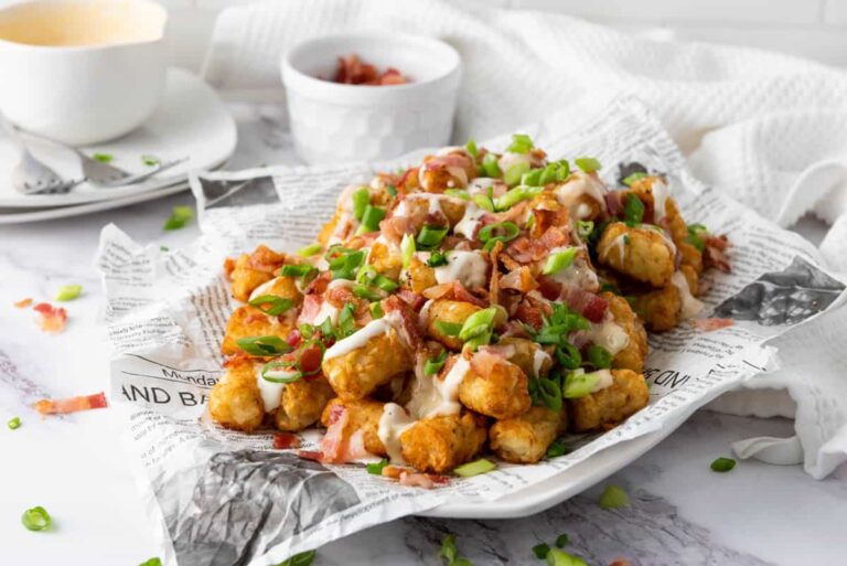 A plate of loaded tater tots topped with bacon, chopped green onions, and drizzled with sauces, served on newspaper-style paper.