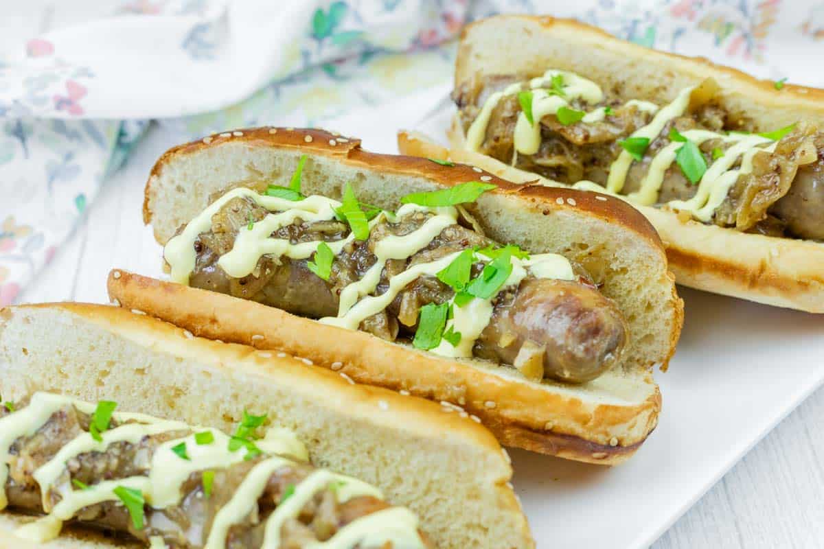 Three Beer Braised Brats in toasted buns topped with sautéed onions, chopped parsley, and a drizzle of yellow sauce, served on a white plate.