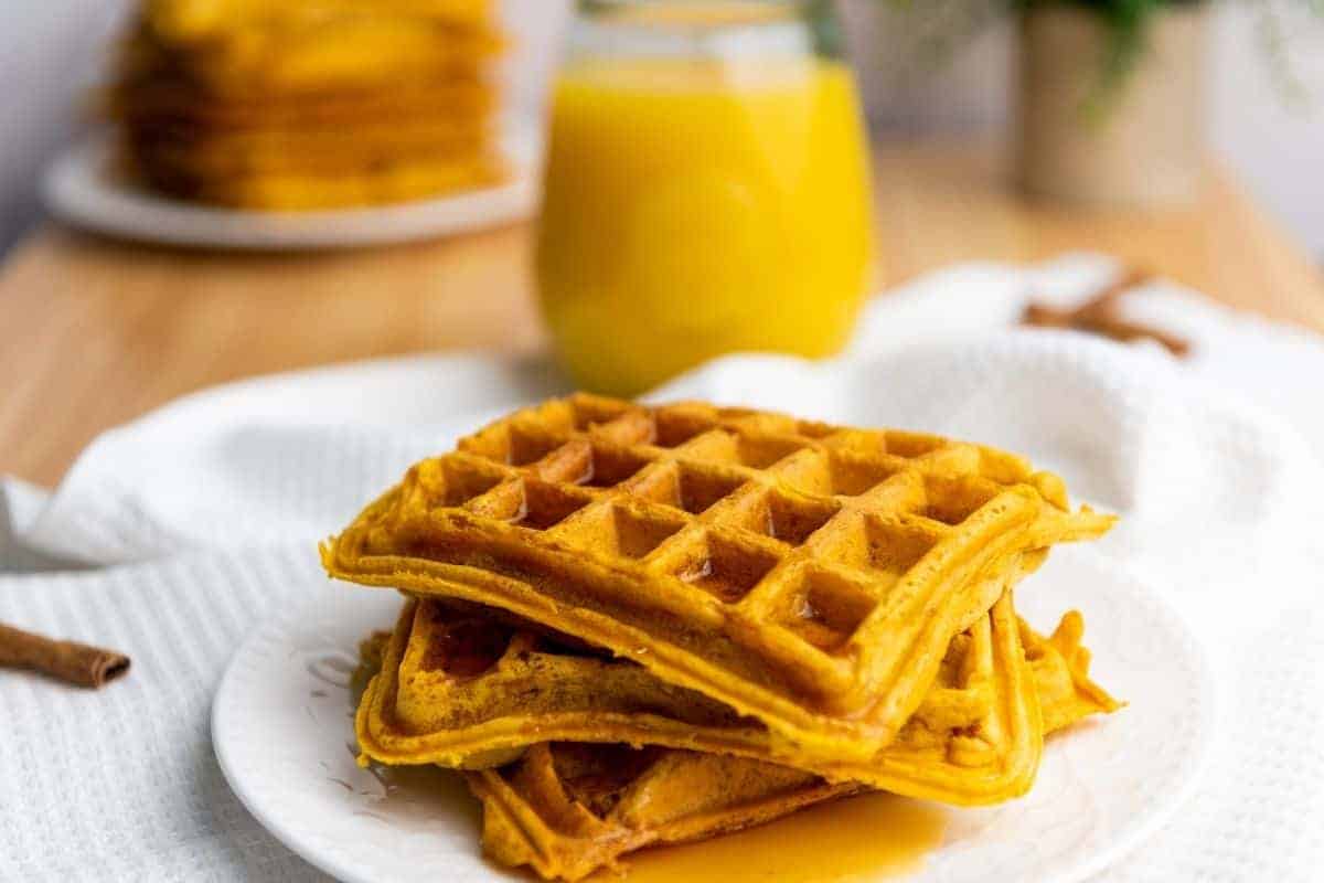 A plate with three stacked pumpkin waffles drizzled with syrup, with a glass of orange juice and more waffles in the background.
