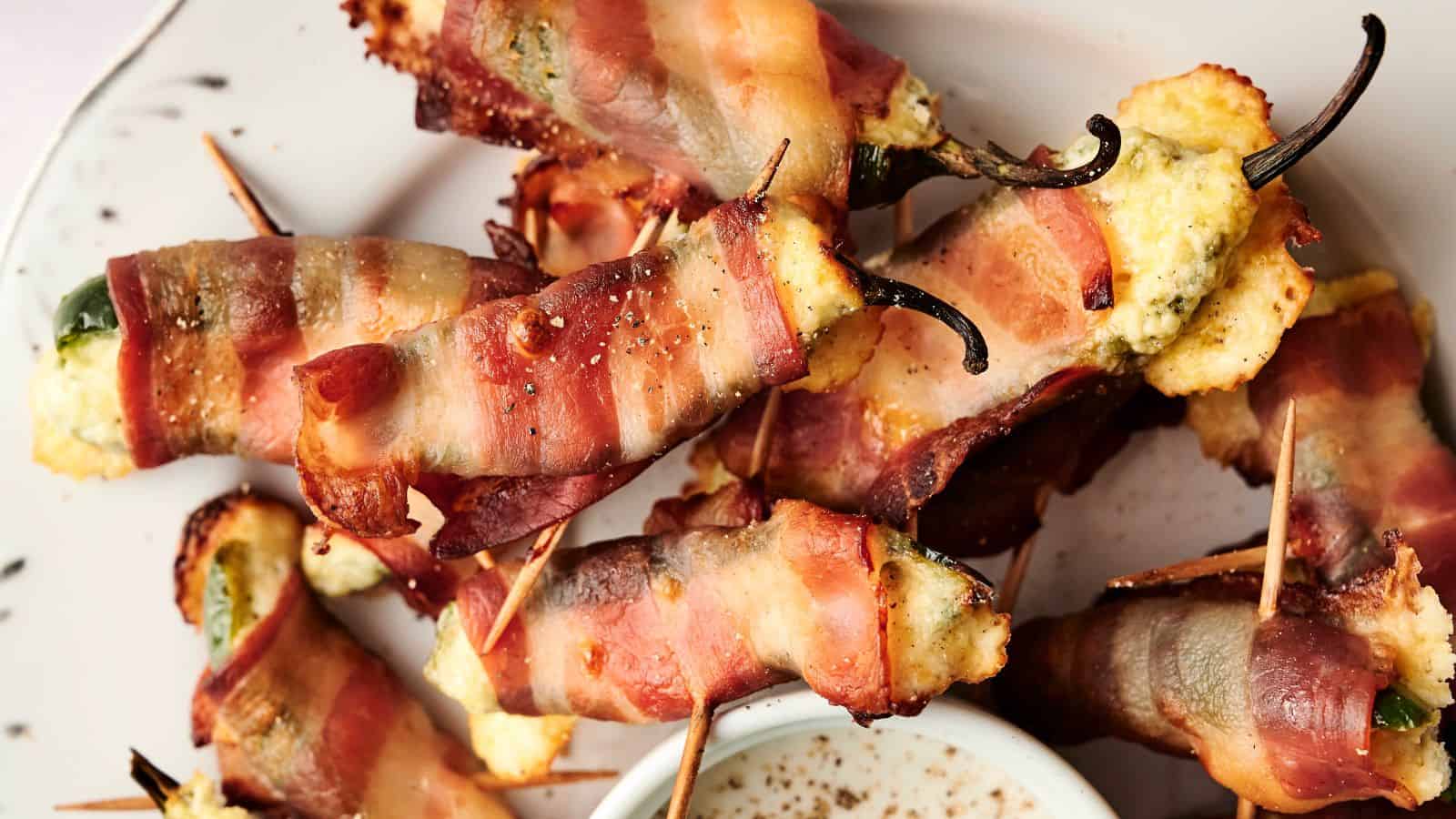 A plate of bacon-wrapped jalapeño poppers filled with cheese, secured with toothpicks, next to a small dish of dipping sauce.