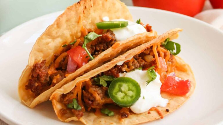 Two ground beef tacos in crispy shells, topped with sliced jalapeños, diced tomatoes, shredded cheese, sour cream, and chopped cilantro on a white plate.