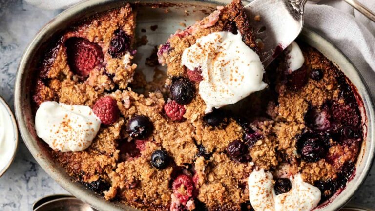 A baked berry crumble topped with dollops of cream and sprinkled with cinnamon, served in an oval dish with a slice being lifted out.