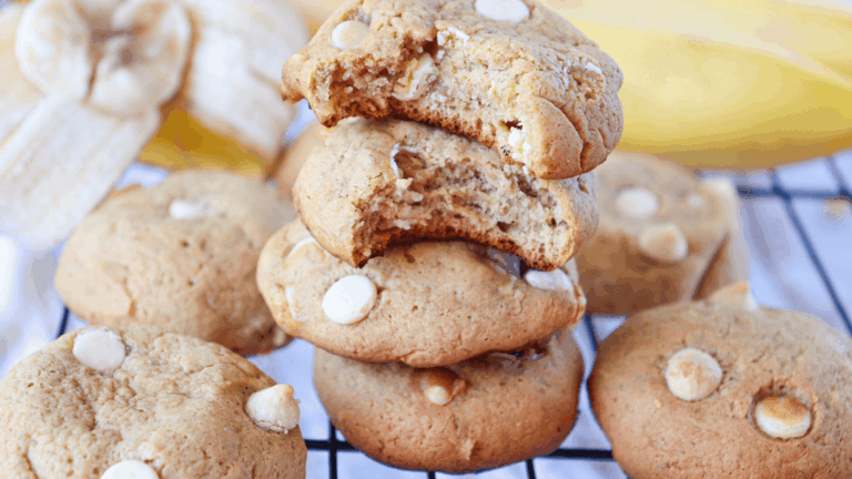 A stack of white chocolate chip cookies, with the top cookie partially bitten, sits on a cooling rack with bananas in the background.