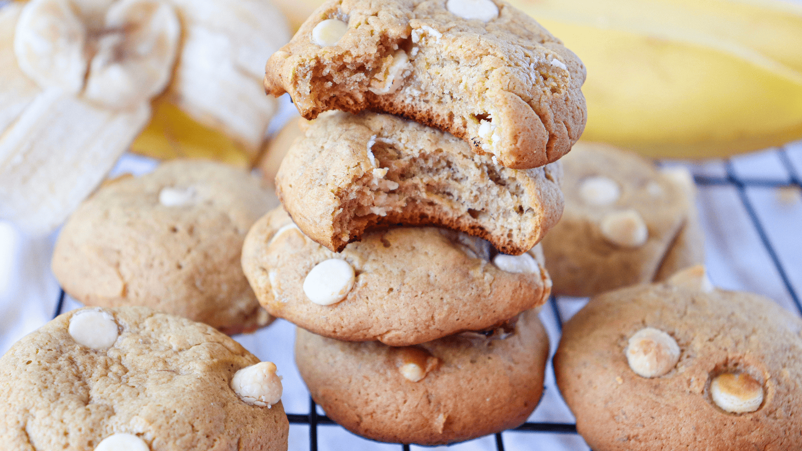 A stack of white chocolate chip cookies with a bite taken out of the top one, placed on a cooling rack with a banana and peeled banana in the background.