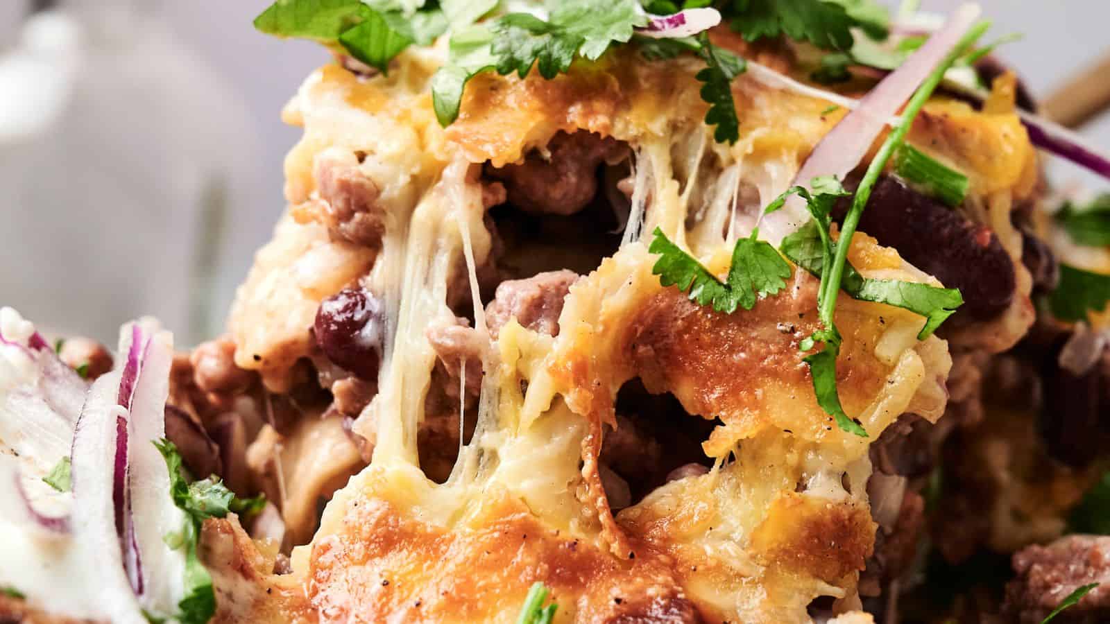 Close-up of a cheesy dish with melted cheese, beef, beans, and scattered cilantro leaves. Red onion slices are visible in the foreground.