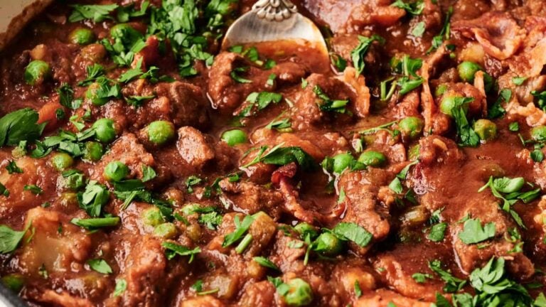 Close-up of a beef stew with green peas, chopped parsley, and a tomato-based sauce, with a spoon partially visible.