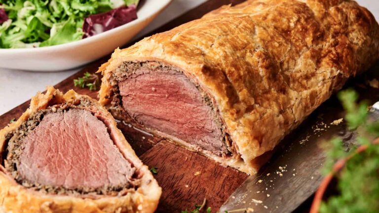 A sliced Beef Wellington shows medium-rare beef wrapped in mushroom duxelles and golden puff pastry on a wooden cutting board, with a green salad in the background.