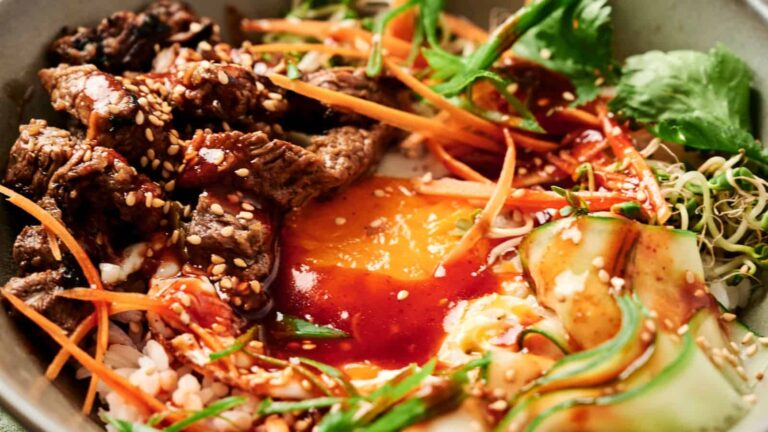 A bowl with grilled beef, shredded carrots, cucumber ribbons, sprouts, rice, fresh greens, a raw egg yolk, and a drizzle of reddish sauce, topped with sesame seeds.