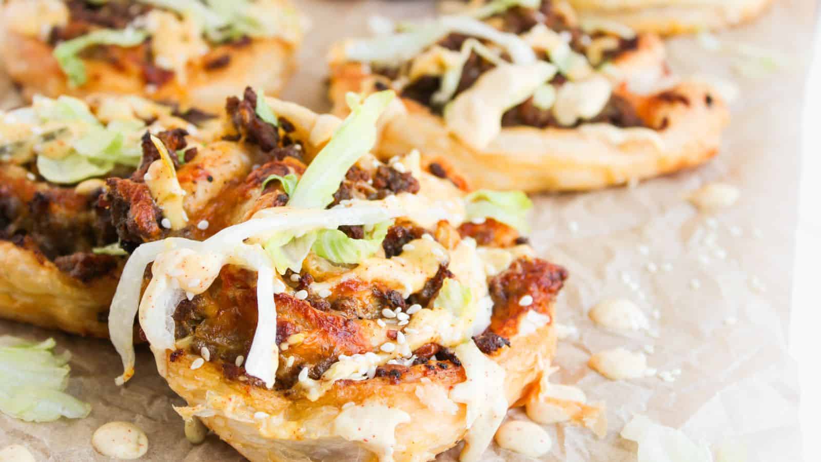 Close-up of puff pastry rounds topped with ground meat, shredded lettuce, sesame seeds, and a creamy sauce, arranged on parchment paper.