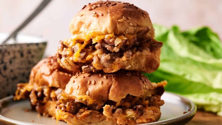 Three sloppy joe sliders with melted cheese are stacked on a plate, with a bowl and lettuce leaves in the background.