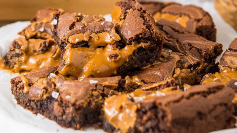 A close-up of fudgy brownies with gooey caramel filling and a sprinkle of coarse salt on top, arranged on parchment paper.