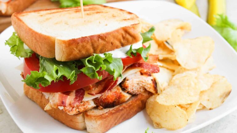 A plate with a sandwich containing grilled chicken, bacon, lettuce, tomato, and mayonnaise on toasted bread, served with potato chips.