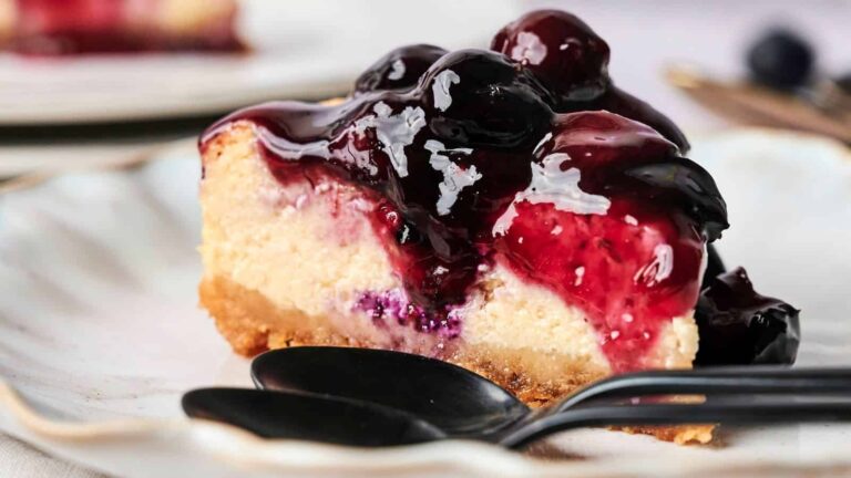A slice of cheesecake topped with glossy blueberry sauce sits on a white plate next to two black spoons.