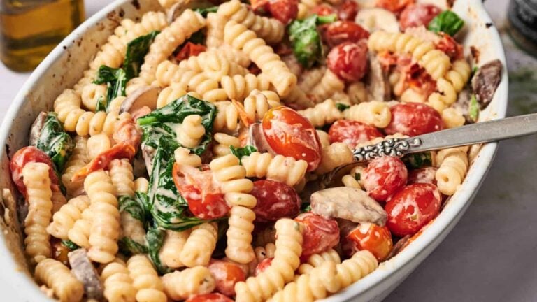 A ceramic dish filled with cooked pasta, cherry tomatoes, spinach, mushrooms, and a creamy sauce, with a spoon resting inside.
