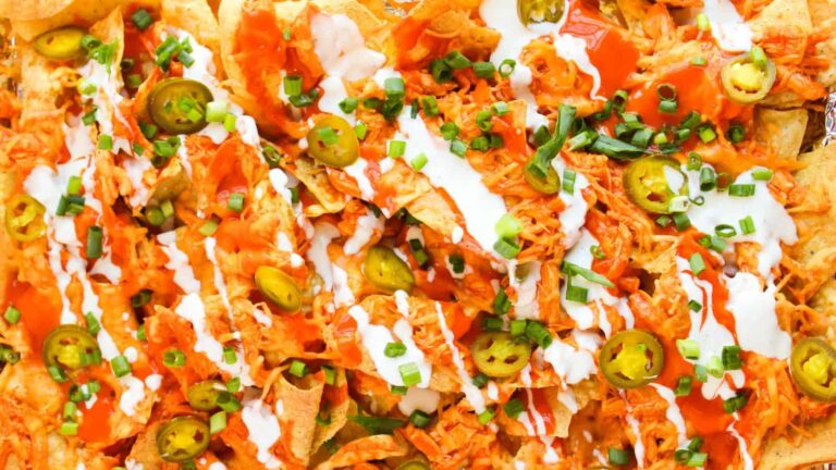 A close-up of nachos topped with shredded chicken, sliced jalapeños, chopped green onions, hot sauce, and drizzles of sour cream.