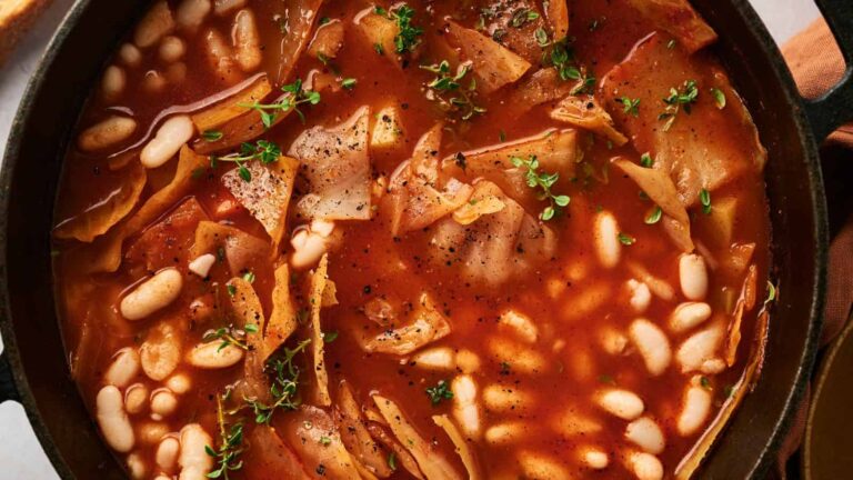 A pot of tomato-based soup with white beans, cabbage pieces, and sprigs of fresh herbs, viewed from above.