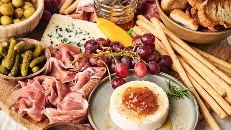 A charcuterie board with prosciutto, cheese, grapes, bread, breadsticks, pickles, olives, and a round of cheese topped with jelly.