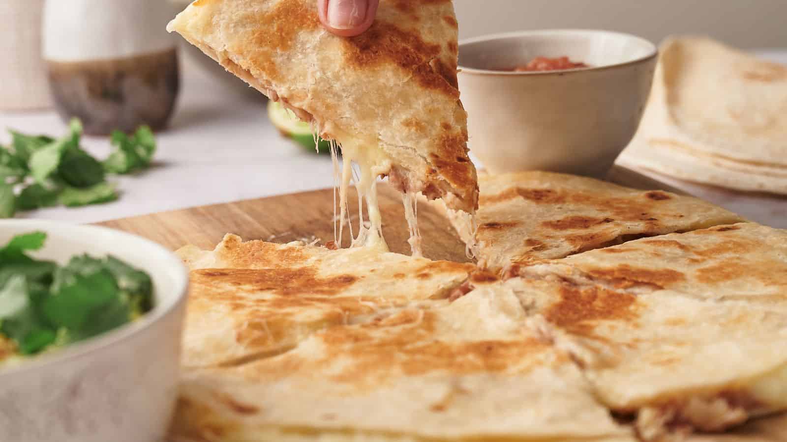 A person lifts a cheesy quesadilla slice from a wooden board. Nearby are bowls of guacamole and salsa on a light-colored table.