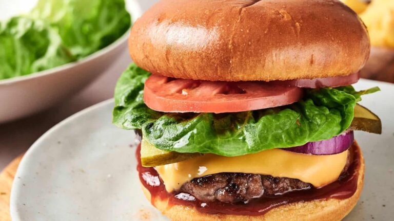 A cheeseburger with lettuce, tomato, pickles, onion, cheese, and sauce on a white plate, with a bowl of lettuce in the background.