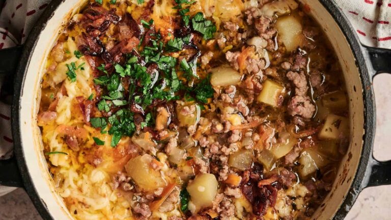 A casserole dish filled with cooked ground meat, diced potatoes, shredded cheese, chopped parsley, and crispy bacon pieces.