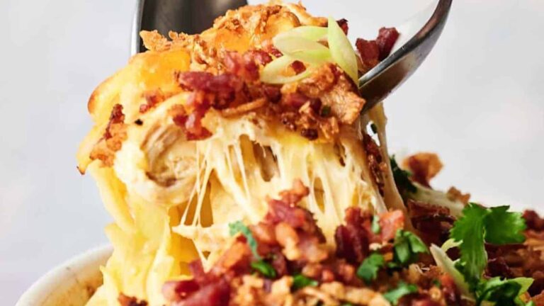 A close-up of a cheesy pasta dish topped with crispy bacon bits, fried onions, and fresh herbs, with a fork lifting a portion.