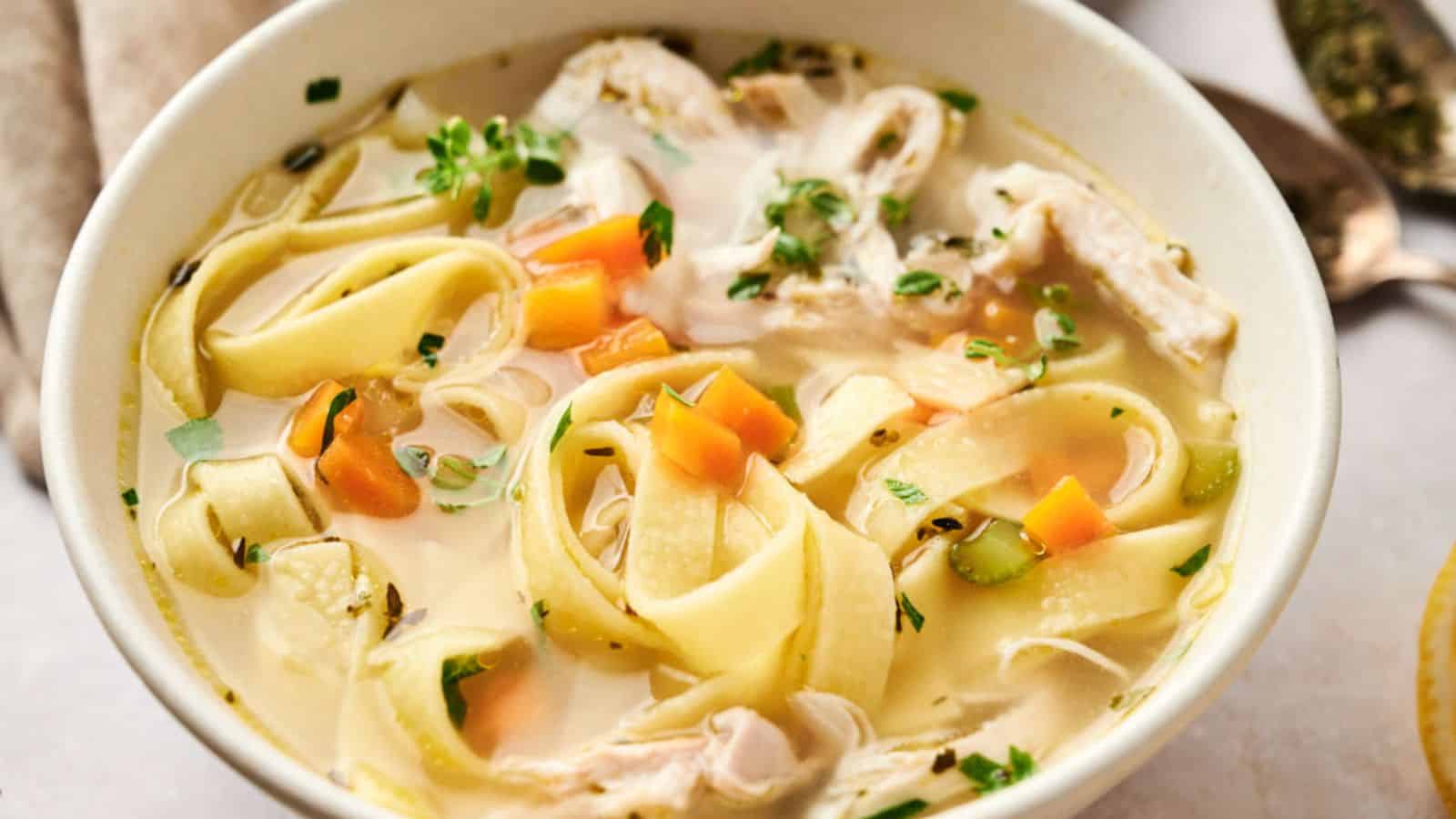 A bowl of chicken noodle soup with wide noodles, chunks of chicken, carrots, celery, and herbs in a clear broth.