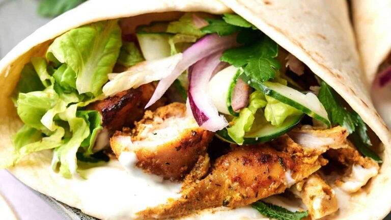 A close-up of a chicken shawarma wrap filled with grilled chicken, lettuce, cucumber, red onion, and sauce in a flatbread.